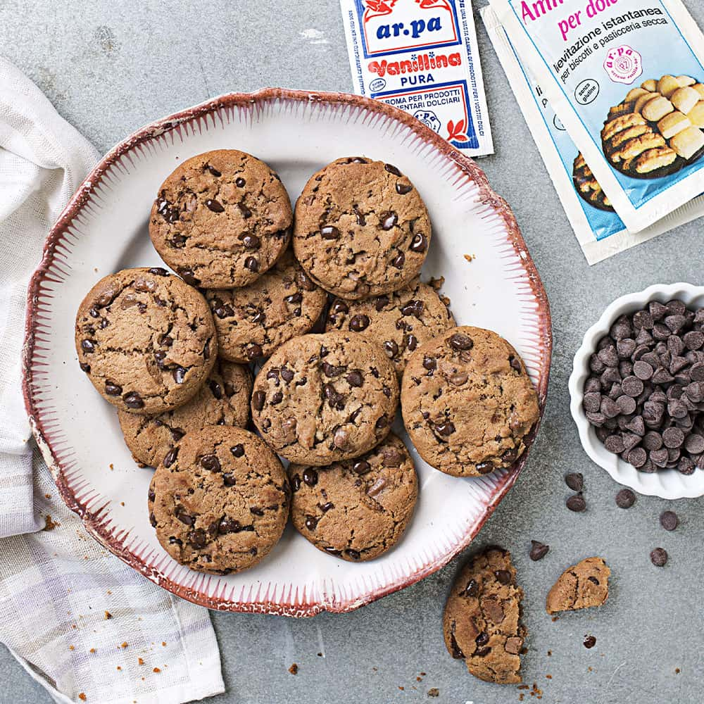 Piatto di biscotti con gocce di cioccolato, insieme a una ciotola di gocce di cacao e due bustine di vanillina e lievito Ar.pa.